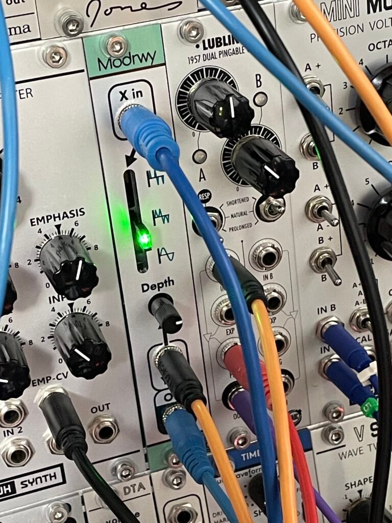 Close-up of a modular synthesizer panel with multiple knobs and colorful patch cables (blue, orange, yellow) plugged into inputs labeled X In, Depth, and Emphasis; a green LED is lit.